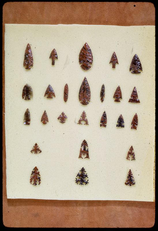 A photographic slide of a series of lithic arrowheads of various designs, materials, and sizes organized in a decorational pattern.