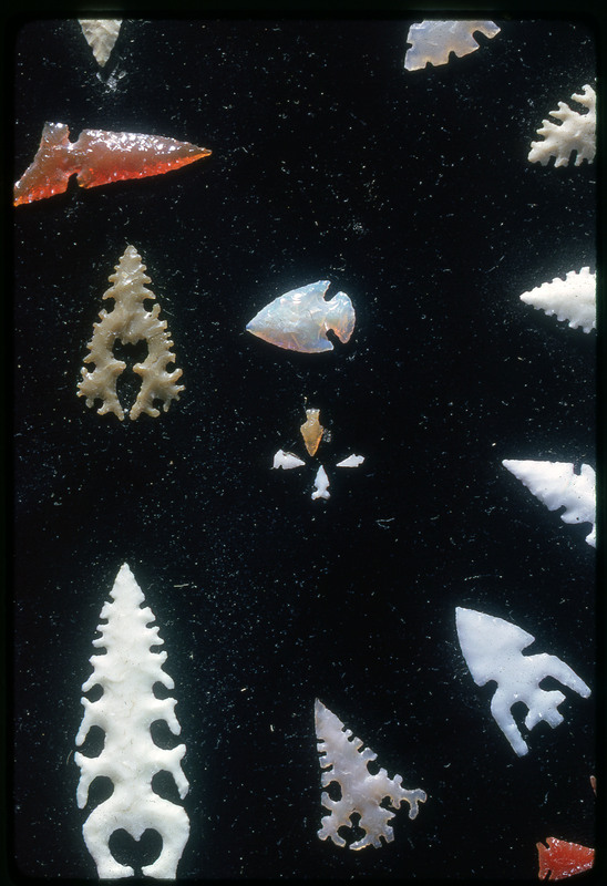 A photographic slide of an artistic shot of lithic arrowheads of various designs, materials, and sizes organized in a decorative concentric pattern.