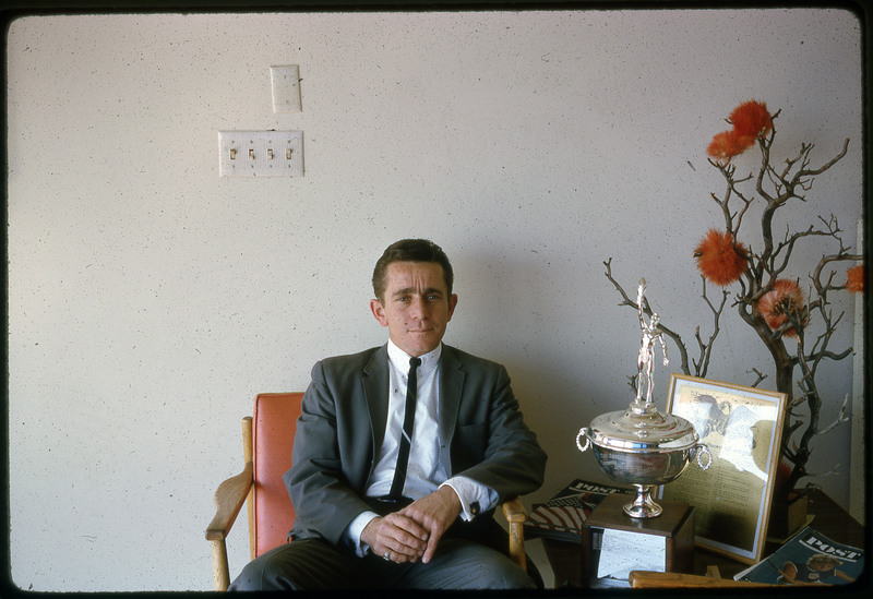 A photographic slide of a man seated in a chair next to a trophy.