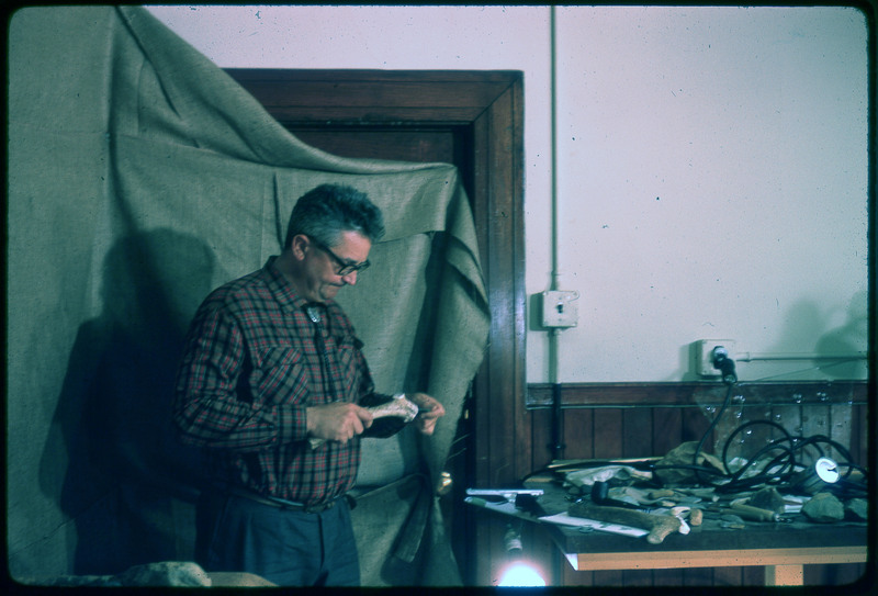 A photographic slide of a man holding a large bone tool used in flintknapping next to a table of assorted flintknapping tools.