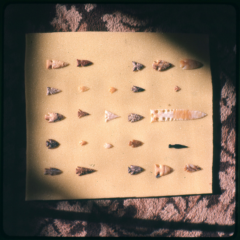 A photographic slide of a series of lithic arrowheads of various designs, materials, and sizes orgnized in a decorational pattern.