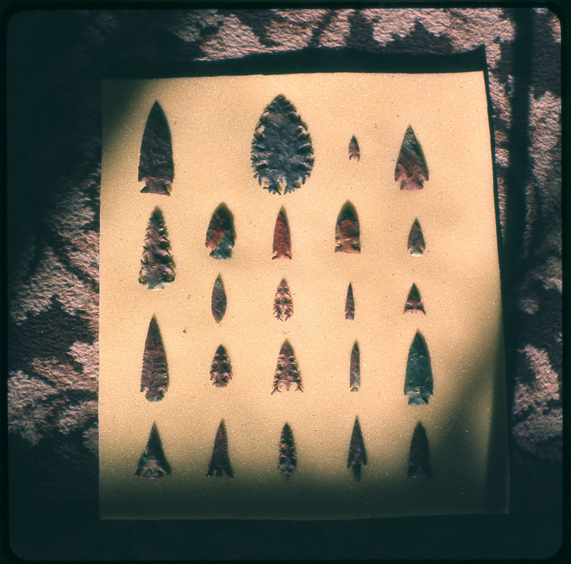 A photographic slide of a series of lithic arrowheads of various designs, materials, and sizes organized in a decorational pattern.