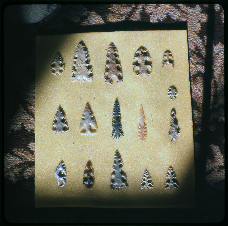 A photographic slide of a series of lithic arrowheads of various designs, materials, and sizes organized in a decorational pattern.