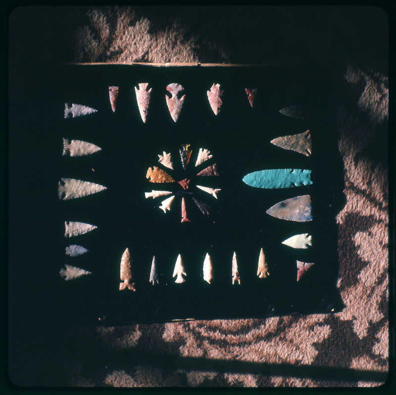 A photographic slide of a series of lithic arrowheads of various designs, materials, and sizes organized in a decorational, concentric pattern.