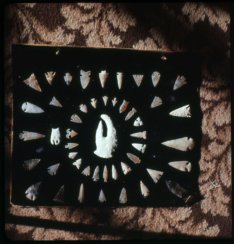 A photographic slide of a series of lithic arrowheads of various designs, materials, and sizes organized in a decorational, concentric pattern.