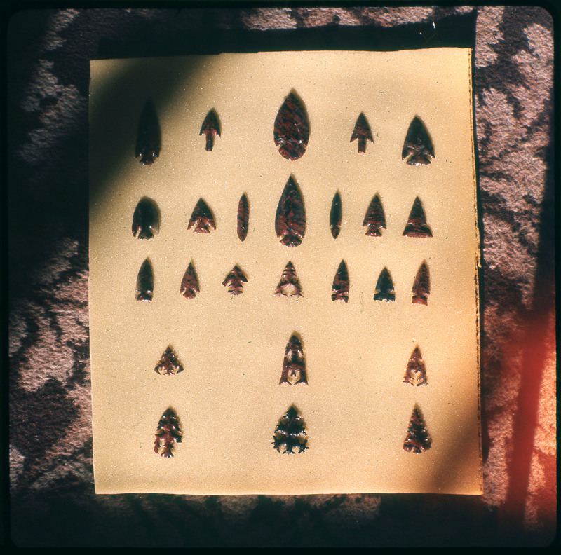 A photographic slide of a series of lithic arrowheads of various designs, materials, and sizes organized in a decorational pattern.