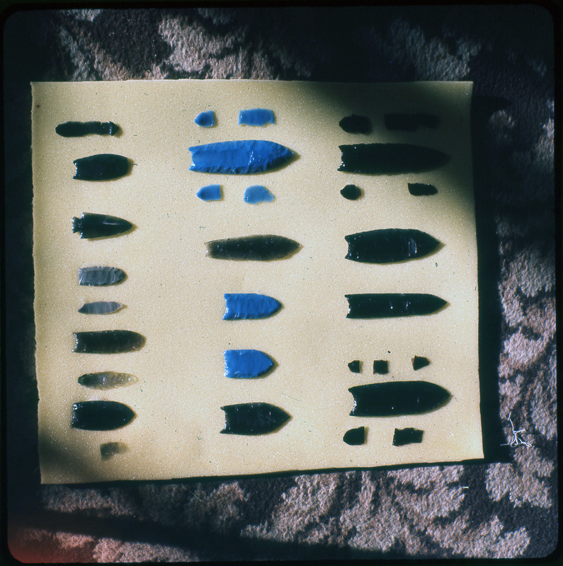 A photographic slide of a series of lithic arrowheads and flakes of various designs, materials, and sizes organized in a decorational pattern.