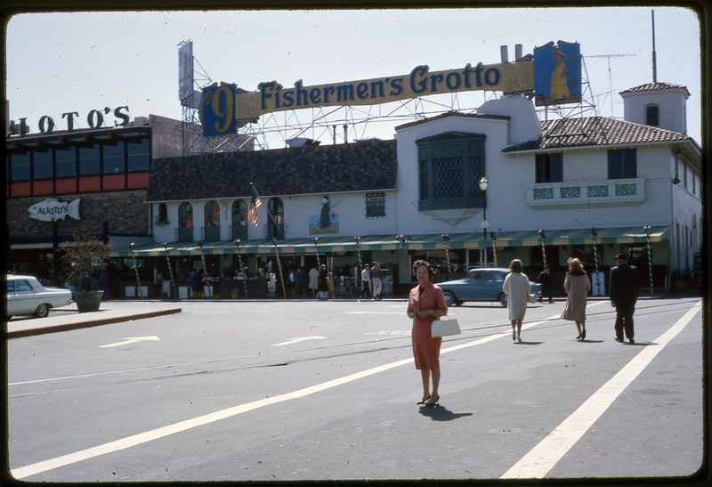 A photographic slide of an urban center with pedestrians, taken in front of a building labelled "Fishermen's Grotto." Evelyn Crabtree stands in the middle of an intersection.