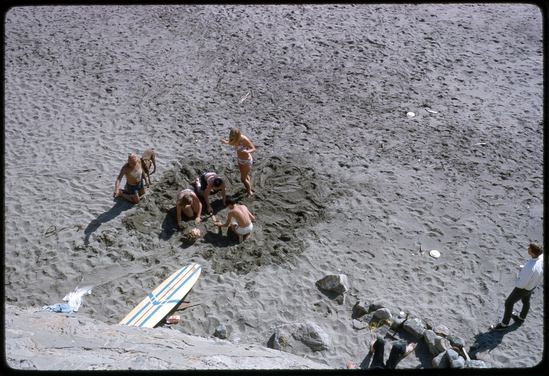 A photographic slide of a group of people digging in the sand together on a beach.