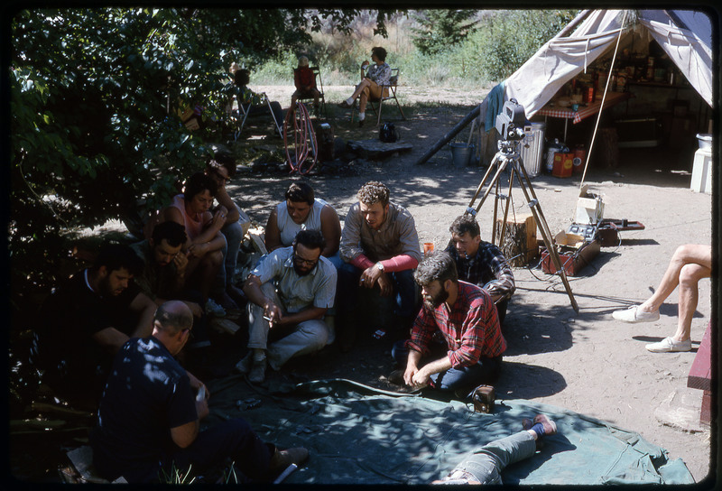 A photographic slide of a group of people huddled together watching as a demonstration is being filmed.