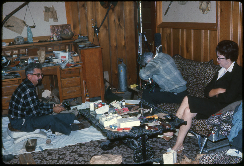 A photographic slide of two men and a woman sitting inside a cluttered room, flintknapping.