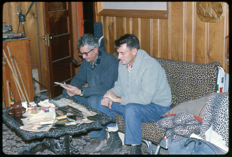 A photographic slide of two men sitting inside on a leapord print couch while flintknapping.