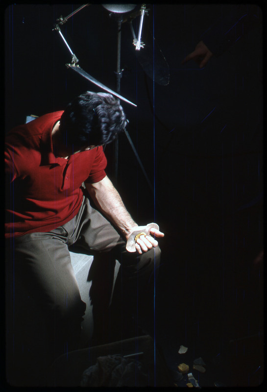 A photographic slide of a man seated while flintknapping under a bright light.