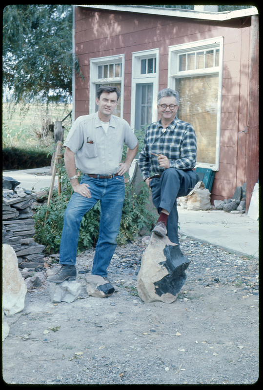 A photographic slide of two men standing in front of a red house, each with a foot resting on a chunk of obsidian.