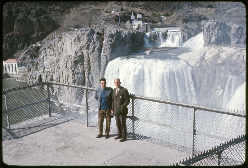 A photographic slide of two men smiling in front of a scenic view of a waterfall.