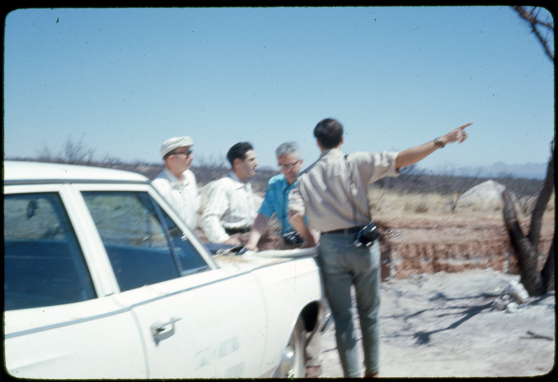 A photographic slide of a blurry view of four men standing over a map on the closed trunk of a car.