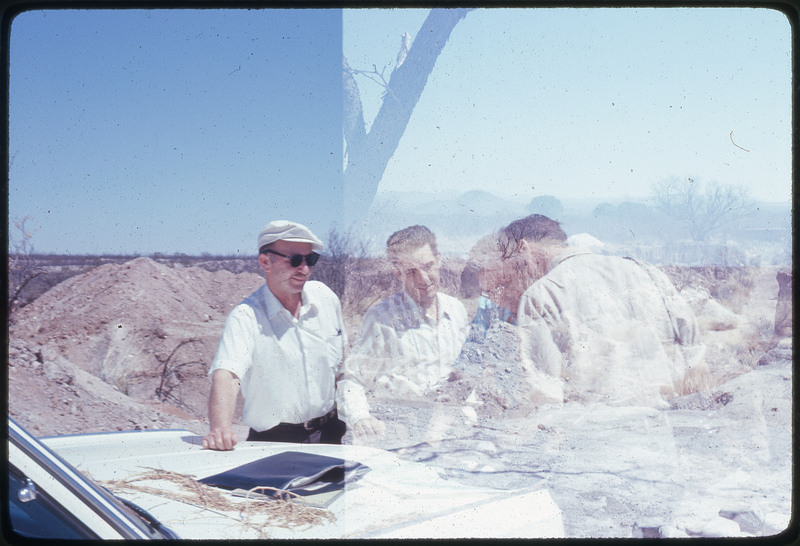 A photographic slide of a superimposed view of four men standing over the trunk of a car. The slide was developed incorrectly, and another image is blurring the original.