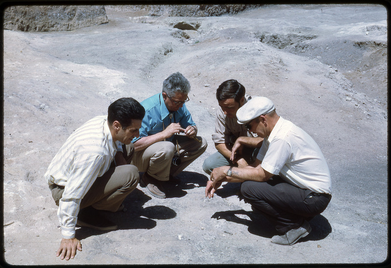 A photographic slide of four men squatting together over rocky ground.