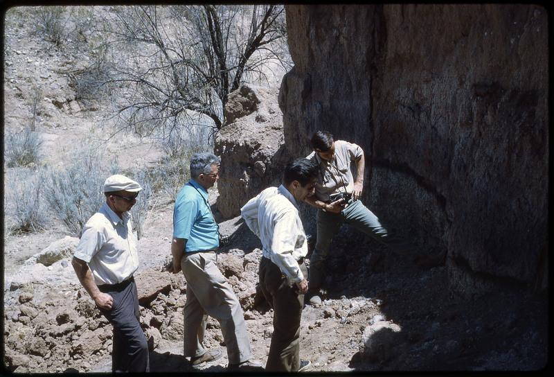 A photographic slide of four men observing the side of a rocky cliff.