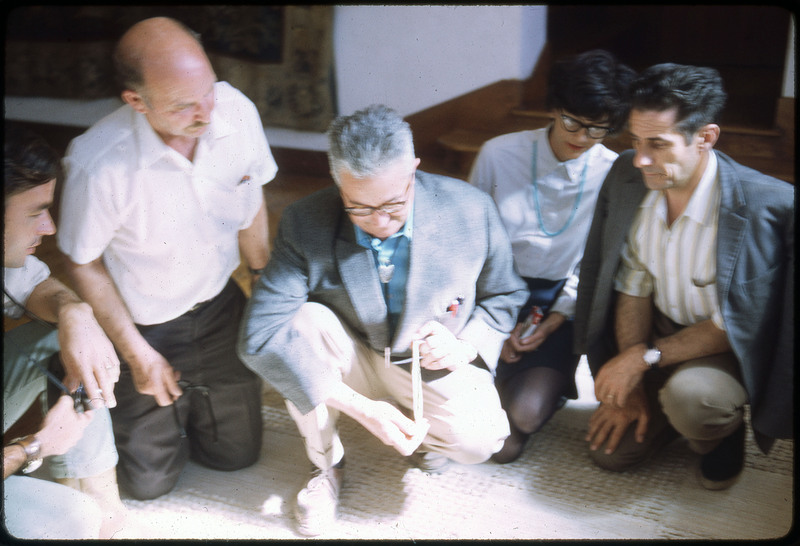 A photographic slide of five people gathered over an artifact that is obscured by bright sunlight.