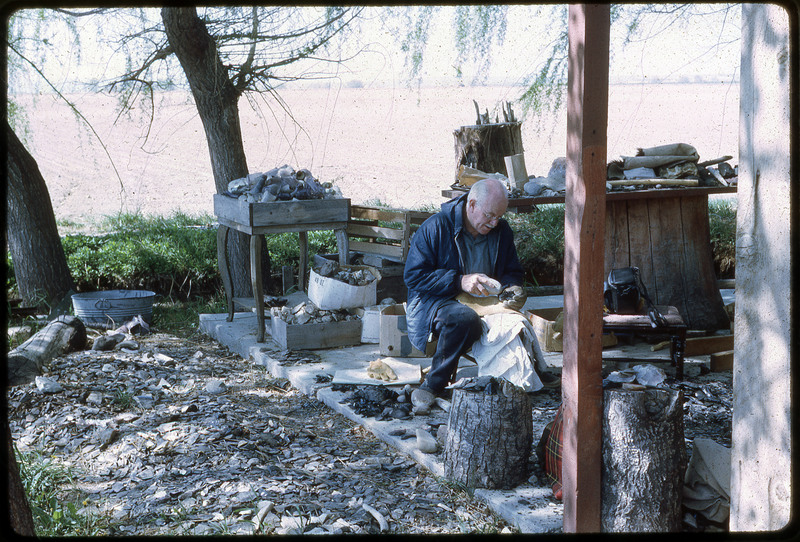 A photographic slide of a man flintknapping on a messy porch.