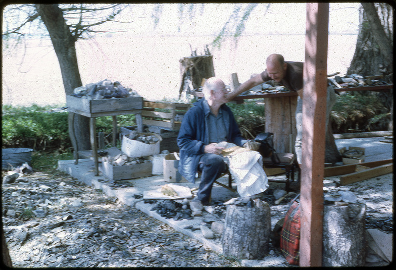 A photographic slide of two men flintknapping on a messy porch.