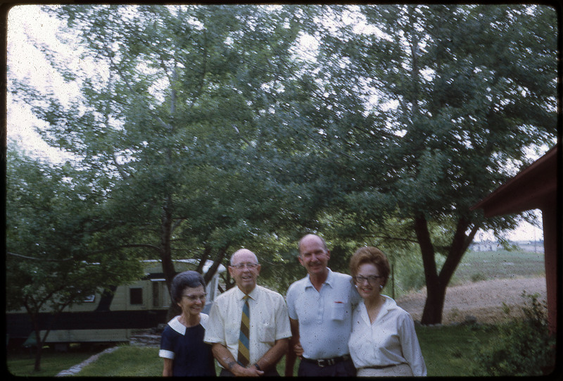 A photographic slide of four people smiling in front of an RV obscured by leafy trees.