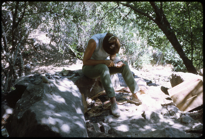 A photographic slide of a person flintknapping under the shade of a tree outside.