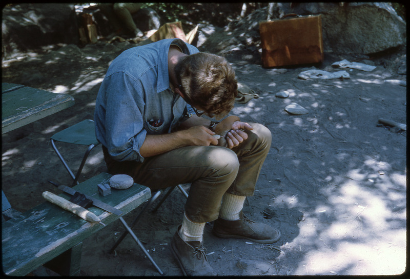 A photographic slide of a person flintknapping while sitting near a park bench outside.