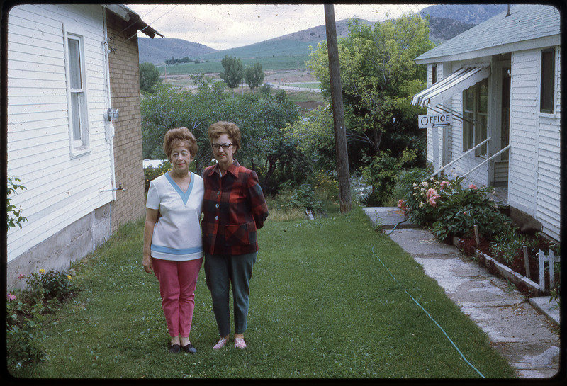 A photographic slide of Evelyn Crabtree and a woman smiling in the yard between two buildings.