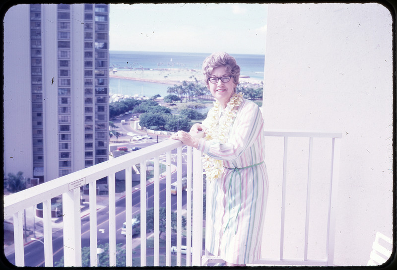 A photographic slide of Evelyn Crabtree smiling on a balcony, wearing a floral lei necklace.