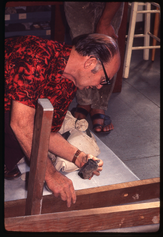 A photographic slide of Don E. Crabtree flintknapping with obsidian while seated inside.