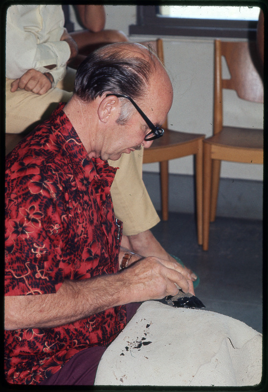 A photographic slide of Don E. Crabtree flintknapping with obsidian while seated inside.