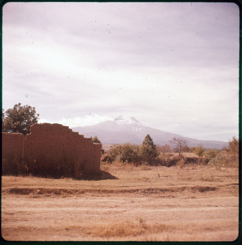 A photographic slide displaying a stone wall in a grassy field. There are a few trees behing the wall, and a large mountain in the background. The entire slide is tinted red.