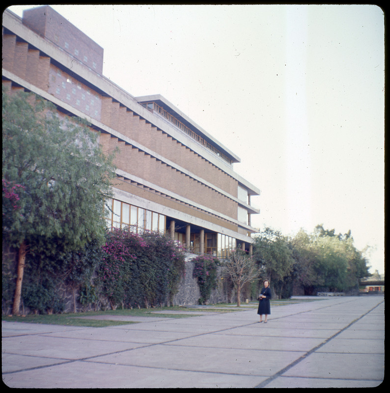 A photographic slide displaying the side of a large building with Evelyn Crabtree in the foreground. The building is surrounded by trees.