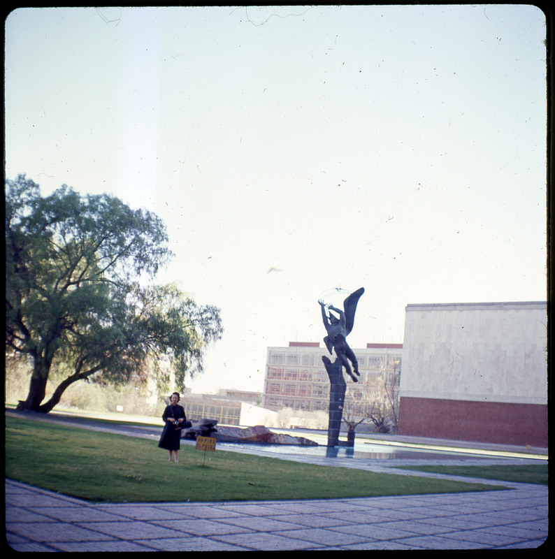 A photographic slide displaying a statue of a winged humanoid with Evelyn Crabtree in the foreground. The statue looks to be a part of a larger structure, but it is cut off by the frame. There is a large tree to the left, and a building to the right.