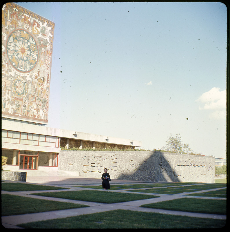 A photographic slide displaying a large decorative mural on the side of a building with Evelyn Crabtree in the foreground. The mural depicts a somewhat geometric scene in faded reds, greens, and browns. There is a wall with similar iconography to its right.