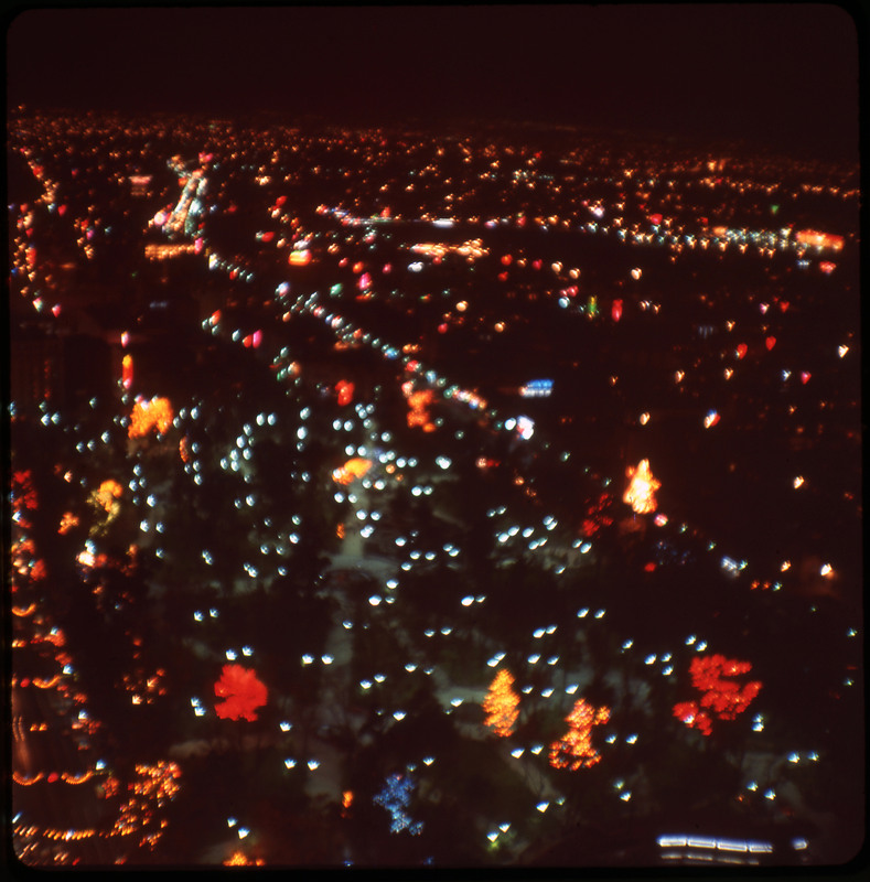 A photographic slide displaying an aerial view of a cityscape at night. Bright, blurry, multicolored lights dot the scene.