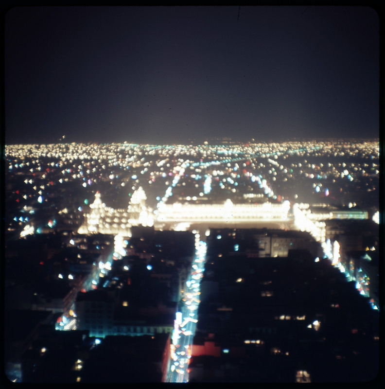 A photographic slide displaying an aerial view of a cityscape at night. Bright, blurry, yellow lights dot the scene.