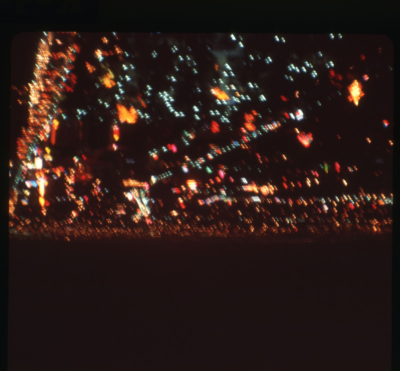 A photographic slide displaying an aerial view of a cityscape at night. Bright, blurry, multicolored lights dot the scene.