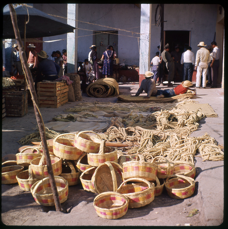 A photographic slide displaying an assortment of woven baskets and many bundles of rope. There are people in the background; sitting, standing, and laying down.