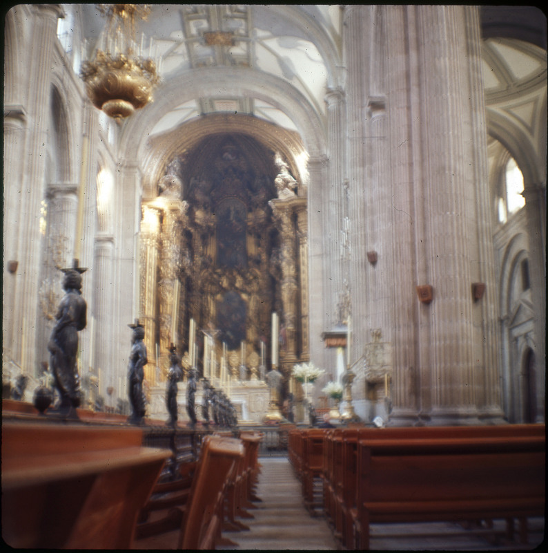 A photographic slide displaying the interior of a cathedral. A grand hall is lined with statues and candles, and there is a golden, ornate structure in the back.