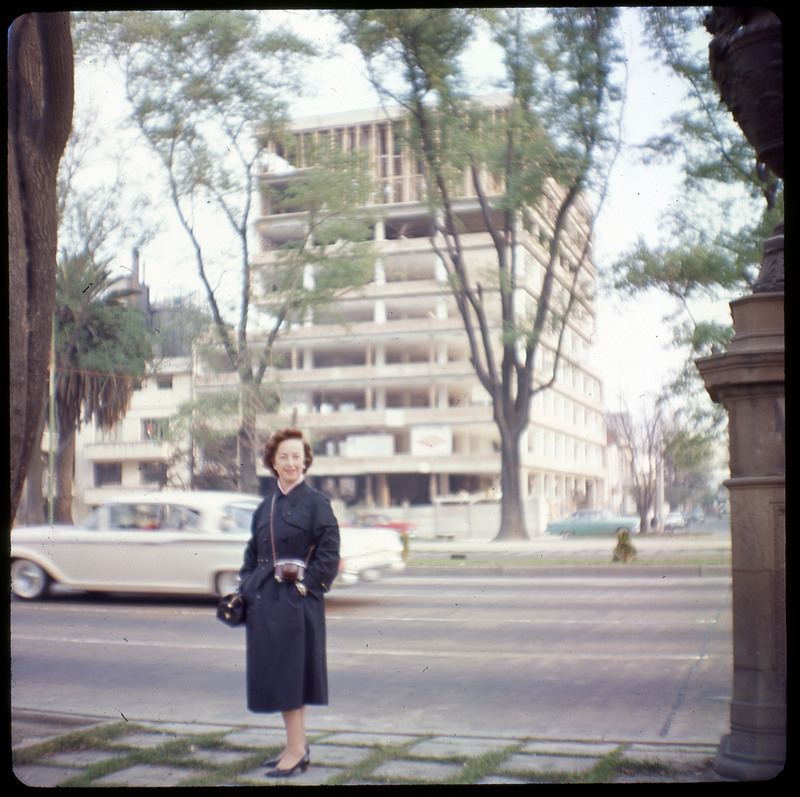 A photographic slide displaying Evelyn Crabtree standing in front of a tall building. The building is primarily columns supporting floors with no walls. There is a car driving on a road between the woman and the building.