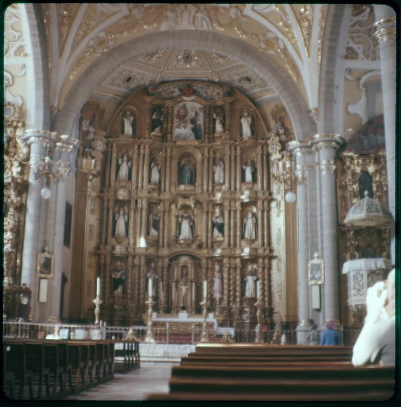 A photographic slide displaying the ornate interior of a cathedral. A large structure is sectioned off into indented windows, each displaying a different statue. The entire slide is tinted red.