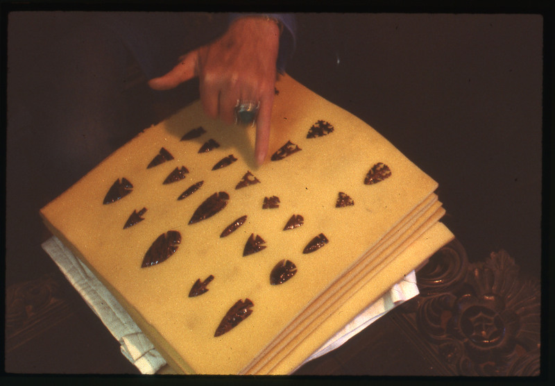 A photographic slide of an arrangement of bifaces and someone pointing to one. The bifaces appear to be arrowheads.