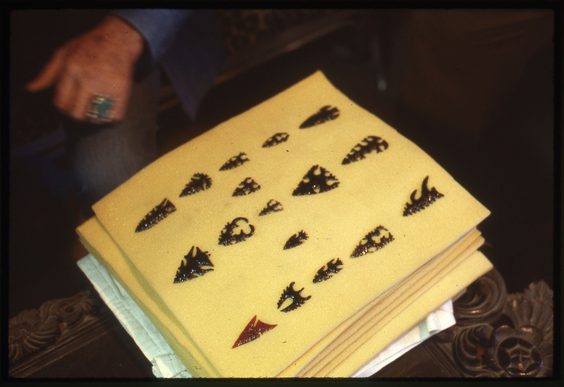 A photographic slide of an arrangement of uniquely flintknapped bifaces and arrowheads. There is a hand in the background.