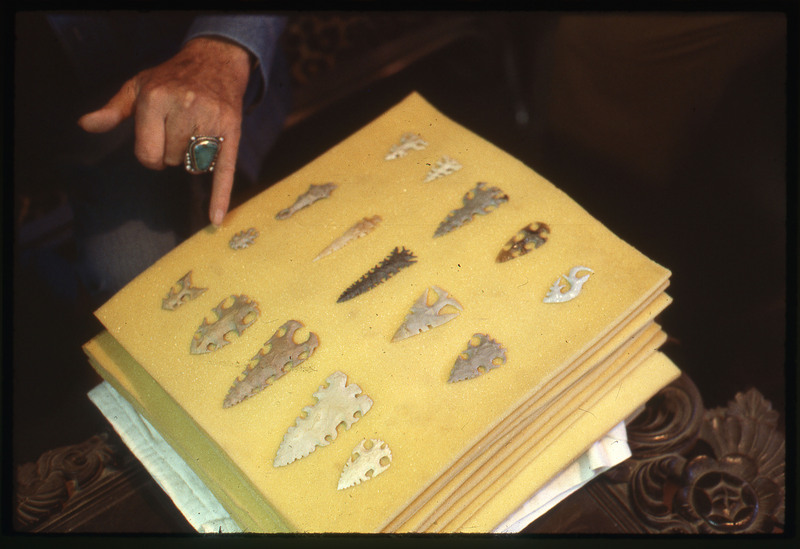 A photographic slide of an arrangement of bifaces. They are made of a white stone and are unique. There is a hand pointing to some of them.