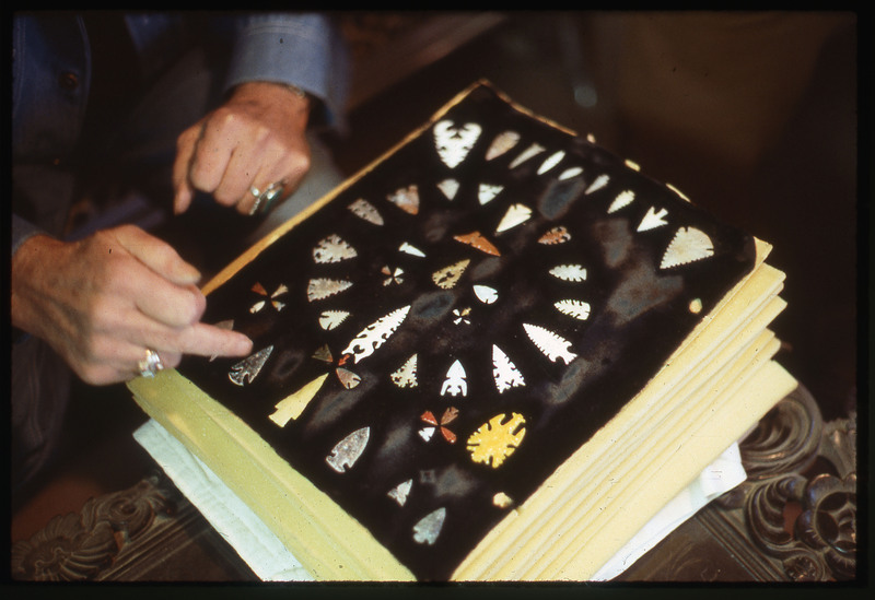 A photographic slide of an arrangement of flintknapped unique and detailed bifaces. They are arranged in a circular pattern and are all different. There are hands in the photo.