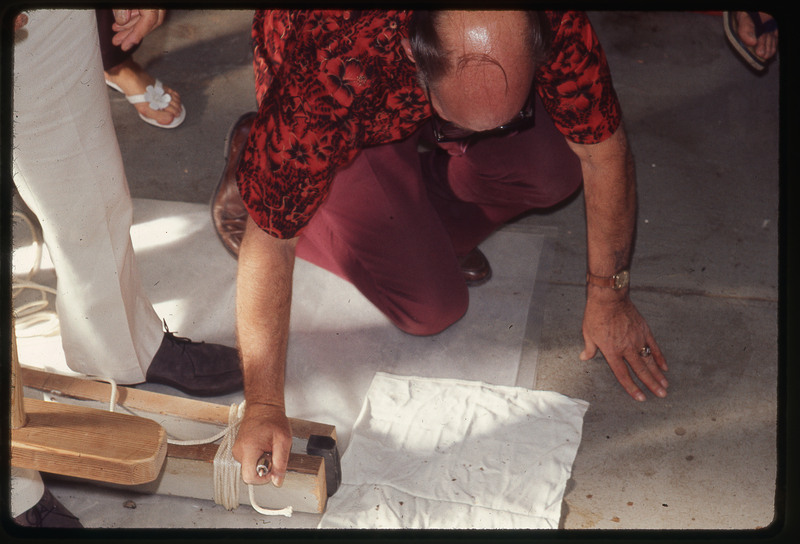 A photographic slide of Donald Crabtree flintknapping using a chest crutch. There are other people in the background.