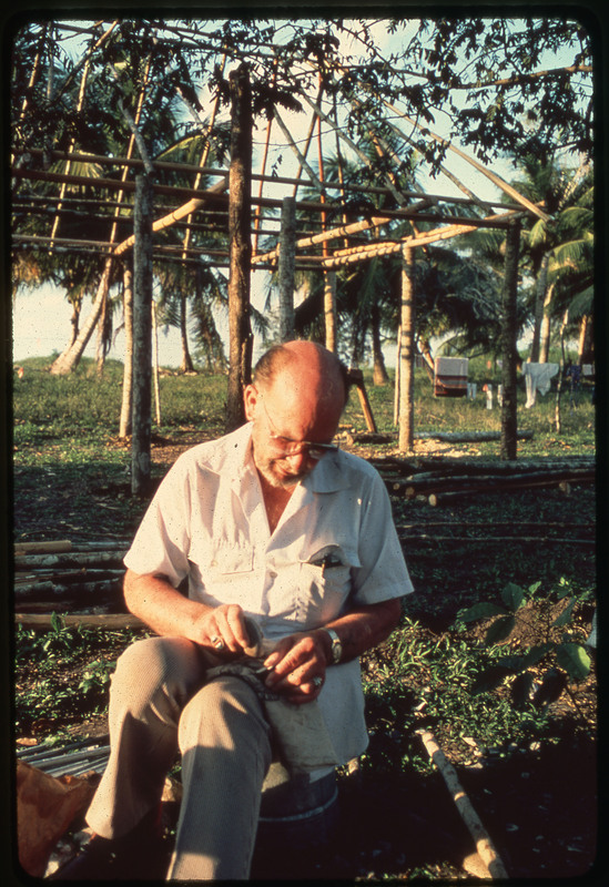 A photographic slide of Donald Crabtree flintknapping in an outdoor space.There is a shelter half built behind him and a clothesline.